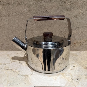 May include: A stainless steel tea kettle with a wooden handle and lid knob. The kettle has a spout and a handle that arches over the top. The polished metal reflects the surrounding environment. A classic kitchenware item.
