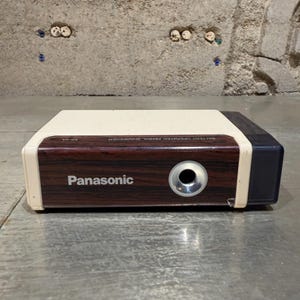May include: A Panasonic KP-2A battery operated pencil sharpener with a cream-colored top, a dark brown wood-grain front panel, and a black rear section. The front features the Panasonic logo, a circular silver-rimmed opening, and text.