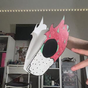 May include: A handmade paper mask featuring a dragon fruit design. The mask has a white base with black dots and a large black eye. The top portion is red with green accents, and the mask is held up by a hand.