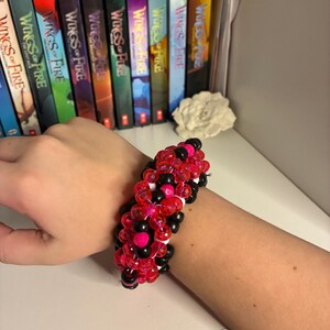 Könnte beinhalten: Ein Perlenarmband mit einer Mischung aus roten, schwarzen, weißen und rosa Perlen. Das Armband wird am Handgelenk getragen. Der Hintergrund zeigt ein Regal mit Büchern und eine dekorative Blume.
