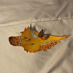 May include: A hand-drawn illustration of a yellow and orange creature's head with a butterfly perched on its back. The creature has a spotted snout, a white stripe, and a closed eye. The butterfly is orange and black.