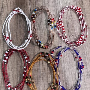 Puede incluir: Seis cuentas de cintura con cuentas de varios colores, incluyendo blanco, rojo, dorado y azul. Las cuentas están ensartadas en múltiples hebras, algunas con acentos decorativos. Las cuentas están dispuestas sobre una superficie de madera.