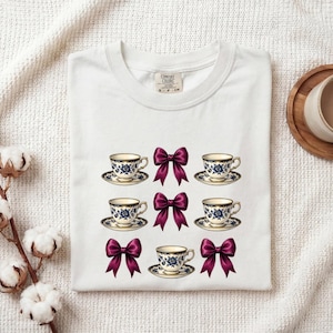 Pode incluir: Uma t-shirt branca com uma impressão gráfica de cinco chávenas de chá com padrões florais azuis sobre pires, dispostas em filas com quatro laços cor-de-rosa escuro intercalados. A t-shirt tem uma etiqueta da marca Comfort Colors no colarinho.