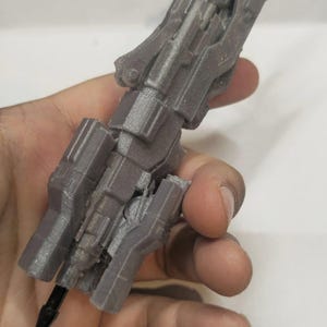Könnte beinhalten: Graues, 3D-gedrucktes Objekt, das einer futuristischen Waffe oder einem Werkzeug ähnelt. Das Objekt hat ein komplexes, eckiges Design mit mehreren Abschnitten und einem Stift, der an der Basis eingesetzt ist. Der Artikel wird in einer Hand vor weißem Hintergrund gehalten.