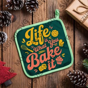 May include: A green square pot holder with the text "Life is What You Bake it" in yellow and orange. The pot holder has a light green border and a loop for hanging. The background includes pine cones, a small red tree, and a wrapped gift.
