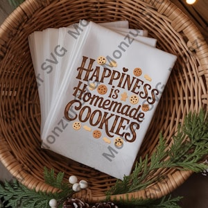 Può includere: Strofinacci da cucina bianchi con la scritta "Happiness is Homemade Cookies" in marrone e illustrazioni di biscotti. Gli strofinacci sono in un cesto di vimini, decorato con verde e pigne.