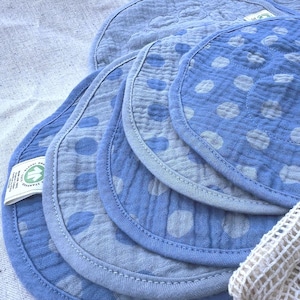 Op de afbeelding: Een stapel ronde, blauwe en lichtblauwe stoffen pads met stippen, gemaakt van gekreukt mousseline materiaal. Elke pad heeft een gestikte rand en een klein wit label met groene tekst en een logo dat Global Organic Textile Standard leest.