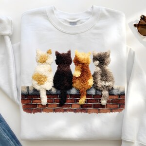Könnte beinhalten: Weißes Sweatshirt mit einem gestickten Design von vier Katzen, die auf einer Backsteinmauer sitzen. Die Katzen sind cremefarben, braun, orange und grau. Die Backsteinmauer ist braun und rot. Das Sweatshirt hat einen Rundhalsausschnitt und lange Ärmel.