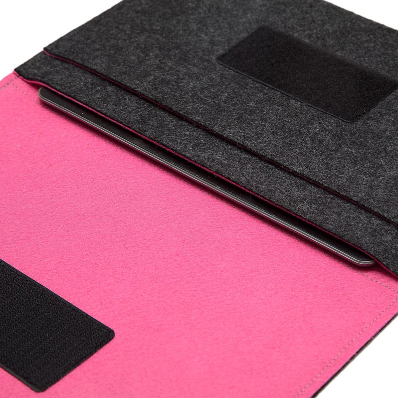 May include: A felt laptop sleeve in dark gray and hot pink. The sleeve is open, revealing the bright pink interior and a laptop. Black velcro closures secure the flap. The edges are neatly stitched with a pink thread.