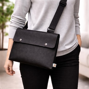 Puede incluir: Bolso mensajero negro con cierre de solapa asegurado por dos remaches plateados. El bolso tiene una correa larga y ajustable de color negro y una pequeña etiqueta en el lateral. El bolso se lleva al hombro.