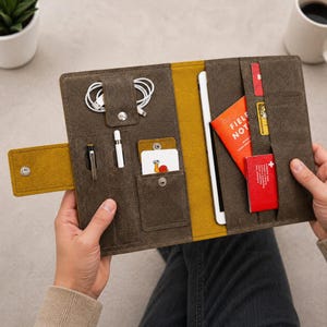 Pode incluir: Capa de tablet em couro castanho com detalhes amarelos. Contém um tablet, cartões e um pequeno caderno com o texto "FIELD NOTES". Inclui também uma caneta, uma caneta stylus e fones de ouvido brancos.