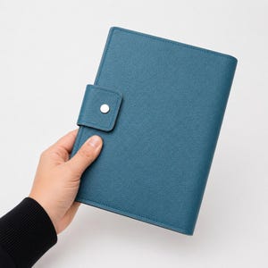 May include: Teal leather-bound journal with a snap closure. The journal is held in a person's hand, showing the textured surface and the silver snap. The cover is a solid teal colour with a stitched border.