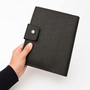May include: Black leather journal with a textured surface and a snap closure. The journal is held in a hand against a white background. The journal has a rectangular shape.
