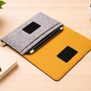 May include: A gray felt laptop sleeve with a black square patch, open to reveal a yellow felt interior. A black stylus is inside. A white mug of coffee, a notebook, and a pen are also on the wooden surface.