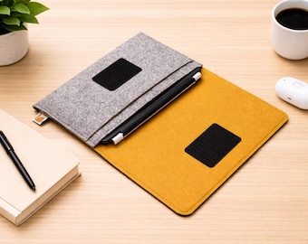 May include: A grey felt laptop sleeve with a black square patch, open to reveal a yellow felt interior. A black stylus is inside. A white mug of coffee, a notebook, and a pen are also on the wooden surface.
