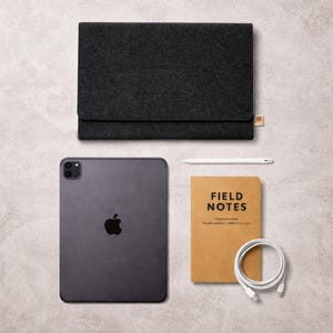 May include: A flat lay featuring a dark gray iPad with a black Apple logo, a black felt sleeve, a white stylus, a brown "Field Notes" notebook, and a white charging cable. The items are arranged on a light gray surface.