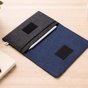 Könnte beinhalten: Eine grau-blaue Filzhülle für ein Tablet oder einen Laptop mit einem weißen Stift darin. Die Hülle hat einen schwarzen quadratischen Aufnäher auf der Vorderseite und ein kleines Etikett. Ein schwarzer Stift und ein Notizbuch befinden sich links. Eine Tasse Kaffee und ein weißes Gerät befinden sich rechts.