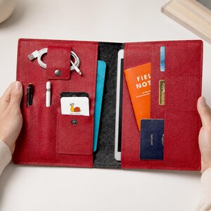 Könnte beinhalten: Rotes Leder-Folio-Organizer mit mehreren Taschen und Fächern. Enthält ein Tablet, ein Notizbuch, Karten, Stifte und ein Ladekabel. Das Folio ist geöffnet und zeigt den Inhalt, gehalten von zwei Händen.