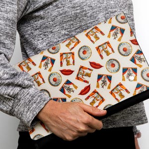 May include: A white clutch bag with a black zipper and a colorful pattern of red lips, eyes, and puppets. The pattern is repeated throughout the bag.