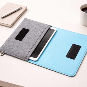 Può includere: Una custodia per tablet in feltro grigio con chiusura nera, che contiene un tablet bianco. La custodia è aperta su una superficie in feltro azzurro. Una tazza bianca di caffè, un taccuino e una penna sono anche sul tavolo.