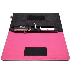 May include: A felt laptop sleeve in black and hot pink, open to reveal a notebook, pen, and smartphone. The sleeve has a black velcro closure and a small tag on the side. The interior is hot pink, and the top flap is dark gray.