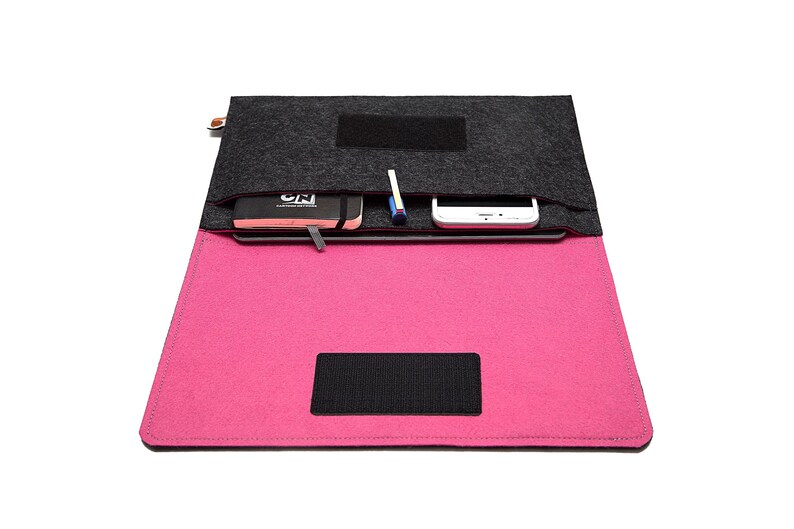 May include: A felt laptop sleeve in black and hot pink, open to reveal a notebook, pen, and smartphone. The sleeve has a black velcro closure and a small tag on the side. The interior is hot pink, and the top flap is dark gray.