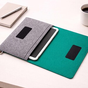 Può includere: Una custodia per tablet in feltro grigio con chiusura in velcro nera, aperta per rivelare un tablet bianco. La custodia poggia su una superficie in feltro verde acqua. Un taccuino, una penna e una tazza di caffè bianca sono anche sul tavolo.