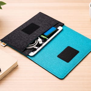 Puede incluir: Una funda de tableta de fieltro gris y turquesa con un parche de velcro negro. La funda está abierta, revelando una tableta, cables de carga y un dispositivo azul. Una taza de café blanca, un pequeño dispositivo blanco, un bolígrafo y un cuaderno también están sobre la superficie de madera.
