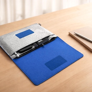 Könnte beinhalten: Eine silber-blaue Laptoptasche mit einem passenden blauen Notizbuch und Stift auf einer Holzoberfläche. Die Tasche ist geöffnet und zeigt ein Telefon und anderes Zubehör. Das Notizbuch ist geschlossen, mit einem silbernen Stift darauf.