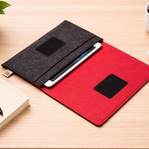 May include: A tablet case made of gray felt, open to reveal a red interior. The case has a black square patch on both the exterior and interior. A tablet is visible inside. A white coffee mug, a mouse, a pen, and a book are on the wooden surface.