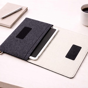 May include: A dark gray felt tablet case with a black rectangular closure, open to reveal a white tablet. A beige notebook with a silver pen and a white mug of coffee are also on the table.