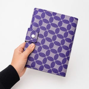 Op de afbeelding: Een paars en grijs patroonboek met een knoopsluiting. De omslag heeft een herhalend ovaal ontwerp in donkerder paars tegen een lichtere grijze achtergrond. Het boek wordt door een persoon vastgehouden.