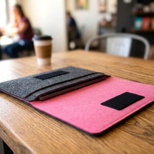 May include: A pink and gray felt laptop sleeve with a black velcro closure. The sleeve is open, revealing a laptop inside. The sleeve is on a wooden table in a cafe setting.