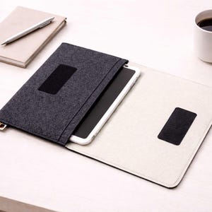 May include: A dark gray felt tablet case with a black closure, open to reveal a white tablet. A white mug of coffee, a notebook, and a pen are on a white surface. The case has a black rectangular patch.