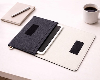 May include: A dark grey felt tablet case with a black closure, open to reveal a white tablet. A white mug of coffee, a notebook, and a pen are on a white surface. The case has a black rectangular patch.