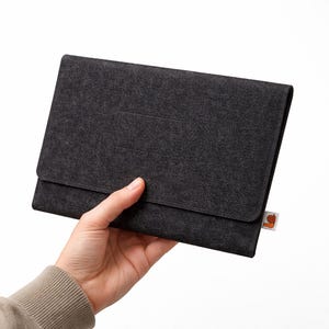May include: A dark gray felt tablet case with a flap closure. The case is held in a hand, showcasing its texture and design. A small tag with a cartoon snail is attached to the side.