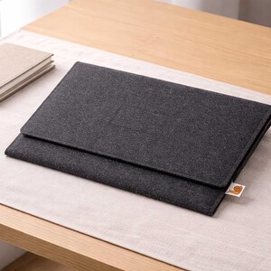 May include: A dark gray felt laptop sleeve with a flap closure, resting on a light beige surface. A small, tan notebook and a gold pen are visible to the left. The sleeve has a small, orange and white tag.
