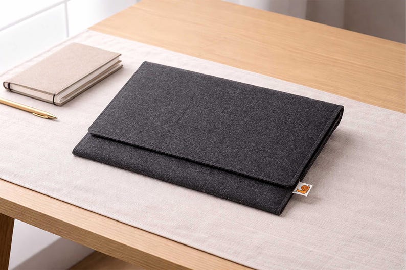 May include: A dark gray felt laptop sleeve with a flap closure, resting on a light beige surface. A small, tan notebook and a gold pen are visible to the left. The sleeve has a small, orange and white tag.