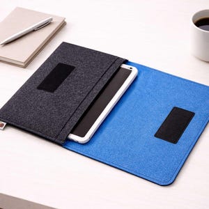May include: A tablet case made of dark gray felt, open to reveal a white tablet. The case is on a blue felt surface. A notebook with a pen and a mug of coffee are also on the table.
