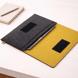 May include: A gray felt laptop sleeve with a black velcro closure, open to reveal a yellow interior. A notebook and pen rest on the table beside the sleeve. The sleeve is designed to protect a laptop or tablet.