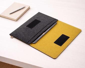 May include: A gray felt laptop sleeve with a black velcro closure, open to reveal a yellow interior. A notebook and pen rest on the table beside the sleeve. The sleeve is designed to protect a laptop or tablet.