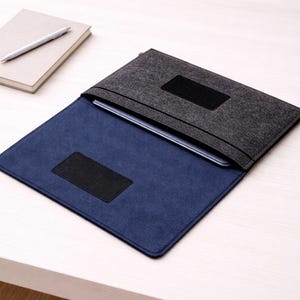 May include: A grey felt laptop sleeve with a navy blue interior, featuring black rectangular patches. A closed beige notebook and a silver pen are on the left, suggesting an office or study setting.
