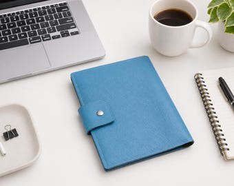May include: A blue leather journal with a button closure sits on a white desk next to a laptop, a cup of coffee, a notepad, and a pen. A small white dish holds a pair of wireless earbuds and a binder clip. A small potted plant is also visible.