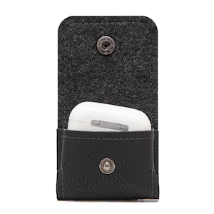 May include: A dark gray felt and black leather case with a snap closure, holding a white wireless earbud charging case. The case is designed to protect and carry earbuds. The case is shown open.