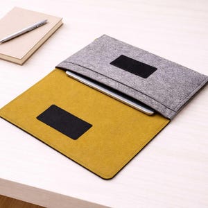 Peut inclure: Une housse de tablette en feutre gris avec un patch rectangulaire noir, ouverte pour révéler une tablette. L'intérieur est jaune moutarde avec un patch noir. Un carnet et un stylo sont en arrière-plan. La housse est conçue pour protéger une tablette.