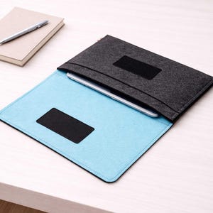 Op de afbeelding: Een grijze vilten laptoptas met een lichtblauwe binnenkant, open om een tablet te onthullen. De hoes heeft een zwarte klittenbandsluiting en een zwarte patch aan de voorkant. Een notitieboek en pen staan op de achtergrond.
