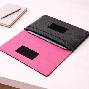 May include: A gray felt tablet sleeve with a black accent, open to reveal a pink interior. A tablet is partially visible inside. A notebook and pen are in the background. The sleeve has a rectangular black patch.