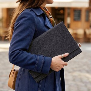 May include: A dark gray felt portfolio held by a person wearing a navy blue coat. The portfolio has a flap closure. A small tan leather purse hangs from the shoulder. The background shows a European street scene.