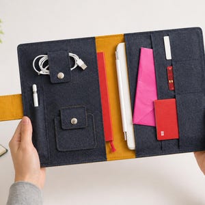 Peut inclure: Un organisateur en feutre bleu marine avec des accents jaunes et orange, contenant une tablette blanche, un stylet blanc et divers accessoires. L'organisateur a plusieurs poches et une fermeture à bouton-pression. Une tasse de café et une petite plante sont en arrière-plan.