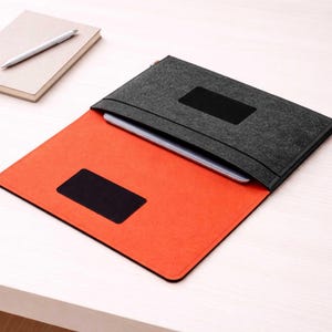 Puede incluir: Una funda gris de fieltro para portátil con un detalle negro y un interior naranja. Un cuaderno y un bolígrafo son visibles en el fondo. La funda está diseñada para proteger un portátil o una tableta.